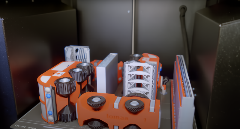 This image depicts a 3D printer with several printed parts on its bed. The parts are intricate, featuring gears, structural supports, and rounded elements with a mix of orange, bla