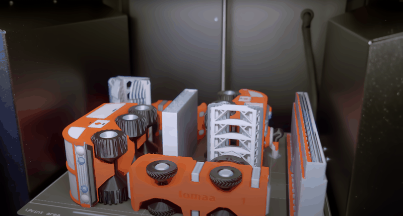 A collection of orange and gray 3D-printed parts within the printing area of the Prusa Core One. The parts, featuring gears and structural components, demonstrate the printer’s ability to produce intricate and functional objects. The setting highlights the machine's operational environment, with parts neatly organized on the print bed.
