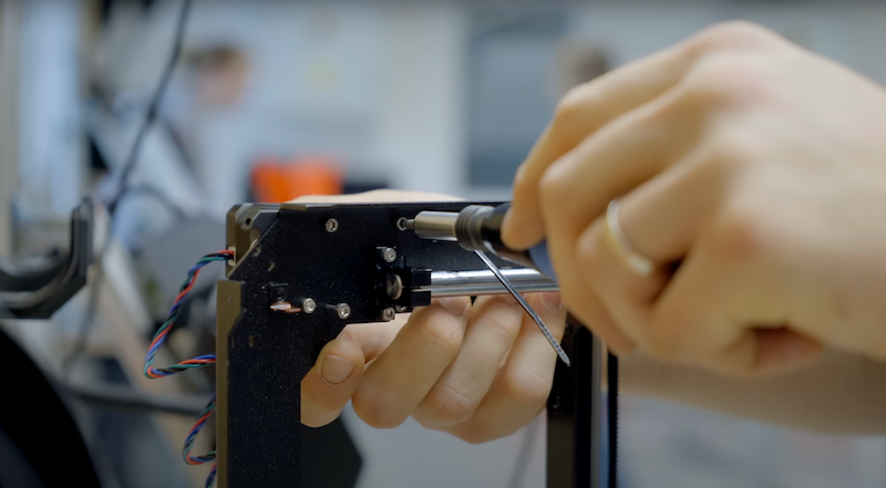 The image shows a close-up of hands using a screwdriver to assemble or maintain a part of a 3D printer. There are wires attached to the structure, indicating an active work in prog