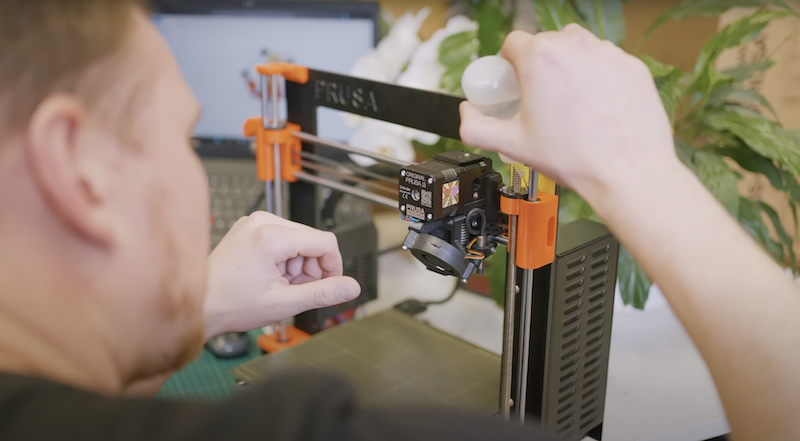 A person is adjusting the 3D printer, featuring the Prusa Core One with distinct orange and black components. The frame displays the Prusa branding. A robust, square frame encloses the print area with visible metal rods and a prominent extruder head. The setting includes a blurred background with a laptop, hinting at a workshop environment with plants for added context.