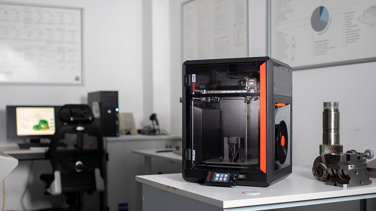 This image displays a 3D printer set up in an office or workshop setting. The printer is idle with no print running. Nearby, there is a desktop computer with a CAD design on the mo