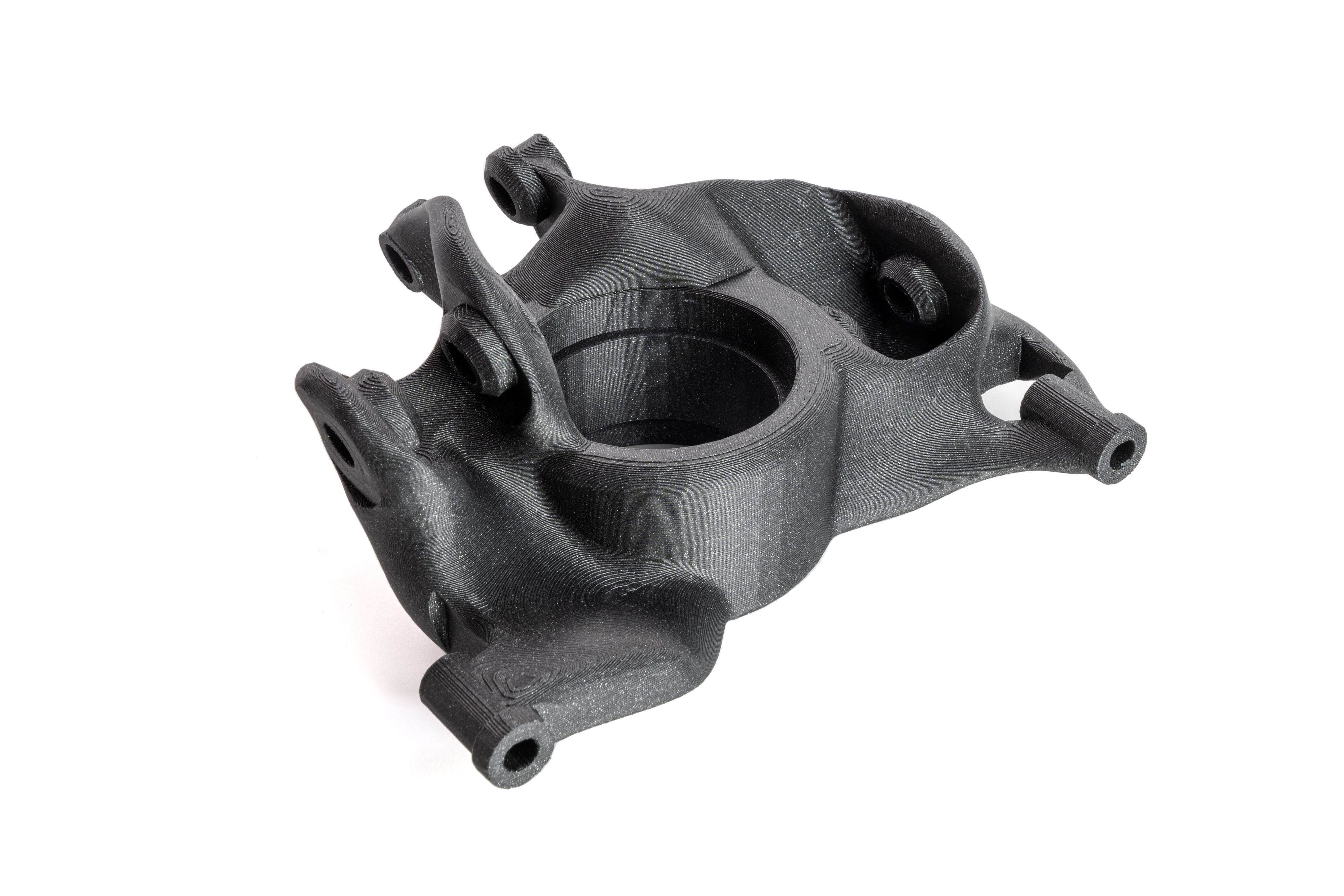 A 3D-printed part in black material. The object has a complex, mechanical design with multiple rounded and angular features, holes, and a large central opening. This is a print sam