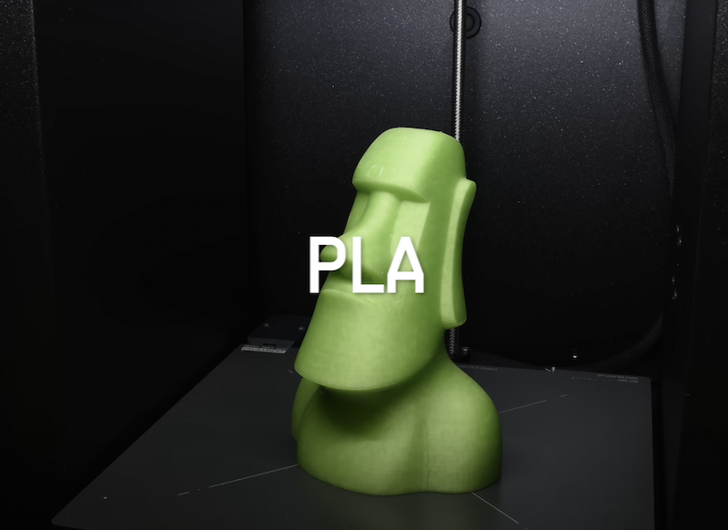A green 3D-printed object resembling a Moai statue is shown inside a 3D printer. The label "PLA" indicates the type of filament used for this print sample.