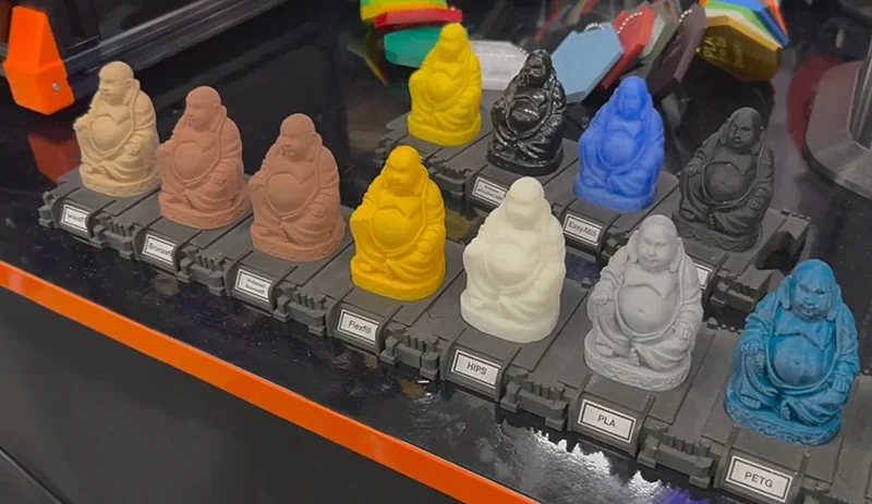 Various colorful Buddha figures, labeled by material type, showcase the Prusa Mini's 3D printing capabilities on a reflective surface.