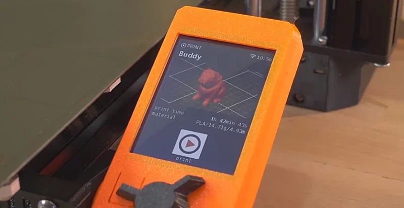 Prusa Mini screen displaying a 3D model print in progress, labeled "Buddy," on a wood surface, featuring an orange frame and print details.