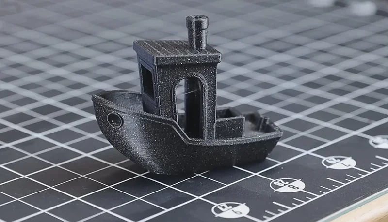 Black 3D-printed boat model on grid mat, created by Prusa Mini printer, highlighting precision and detail.
