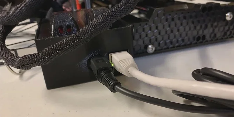 Prusa Mini connections show black and white cables plugged into the 3D printer's black casing with a honeycomb pattern detail, on a table surface.