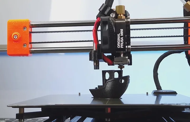 Prusa Mini 3D printer creating a black model boat. Features the printer's nozzle, red cables, and branding in a light background setting.
