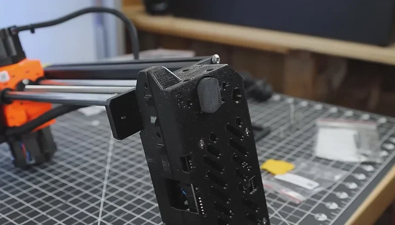 Prusa Mini 3D printer close-up, black and orange frame, on a patterned table with printed parts and tools in a workshop setting.