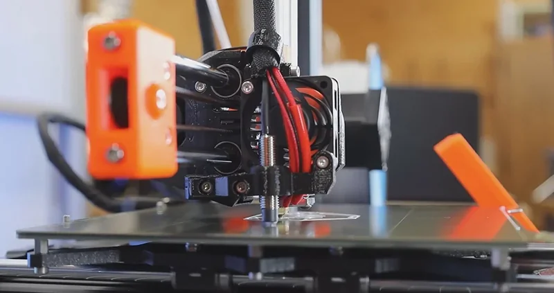 Prusa Mini 3D printer close-up, showcasing its red and black extruder, printing a model on a heated bed, with orange accents in a workshop setting.