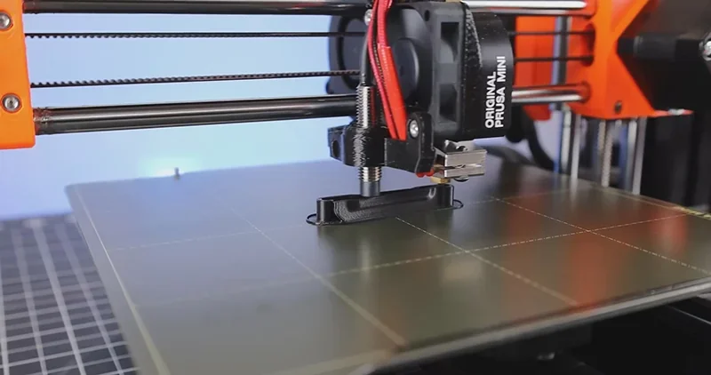 Prusa Mini 3D printer in action, printing a black plastic object on a grid-patterned build plate, showcasing precision and compact design.