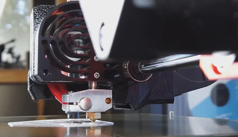 Prusa Mini's print head in action, fabricating a white plastic layer on the build plate, showcasing precision and design within a workshop setting.