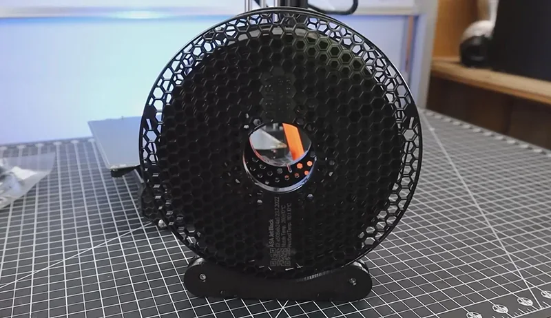 Prusa Mini 3D printer spool holder with black filament on grid-patterned table; hexagonal design, workshop setting.