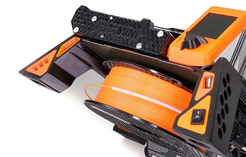Prusa Mini 3D printer close-up showing brand logo, orange filament spool, USB port, and control display, with printed part samples.
