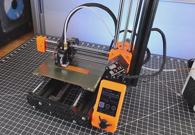3D printer Prusa Mini on a grid table, orange components visible, printing a battery holder; sample created by Prusa Mini shown on bed.