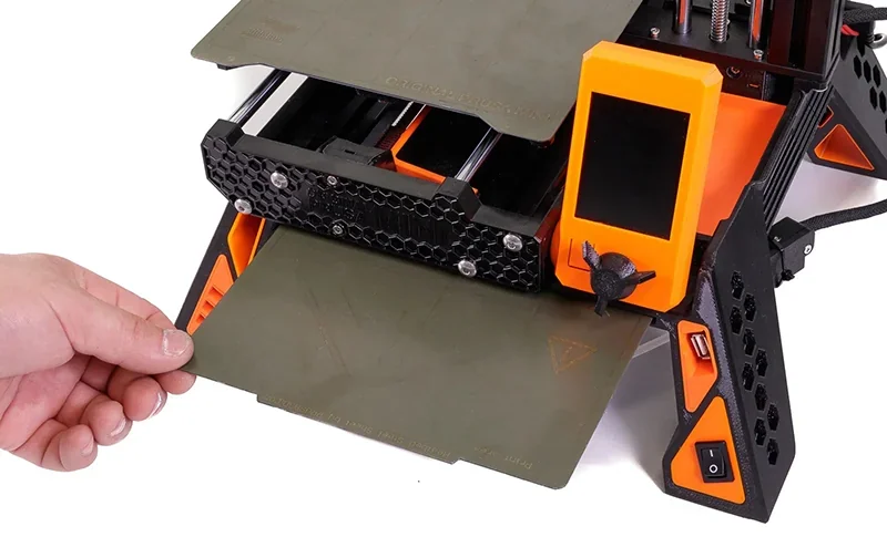 Prusa Mini 3D printer with orange display and honeycomb design, showcasing removable print bed held by a hand, in a close-up view.
