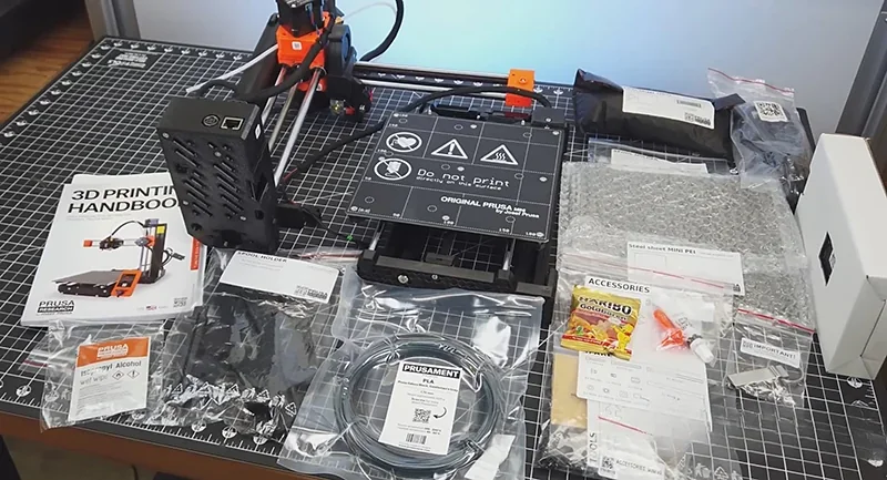 Prusa Mini 3D printer with assembly parts, filament, and accessories displayed on a grid mat with a 3D Printing Handbook.