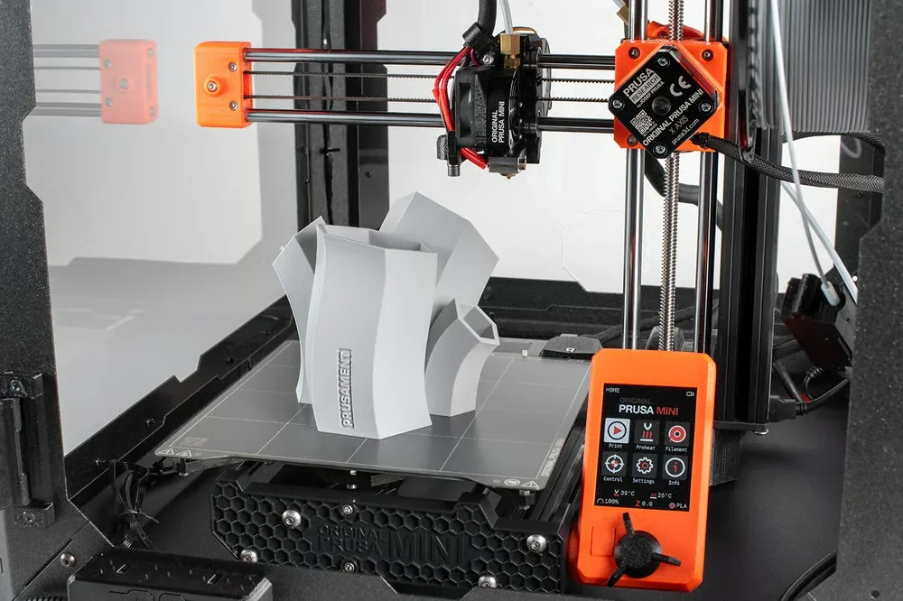 Prusa Mini 3D printer featuring an intricate gray 3D-printed object, with orange accents and a digital display, showcasing precision and design capabilities.
