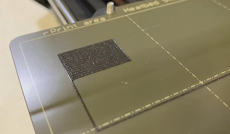 Prusa Mini print bed with a small, dark 3D print sample. Heated print area marked with dashed lines on a green surface.