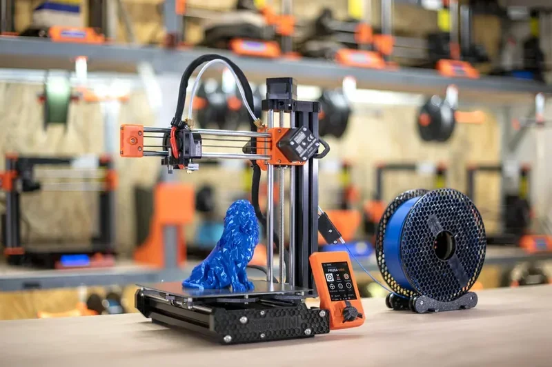 Prusa Mini 3D printer on a desk, printing a blue lion model; visible filament spool and control display in a workshop setting.