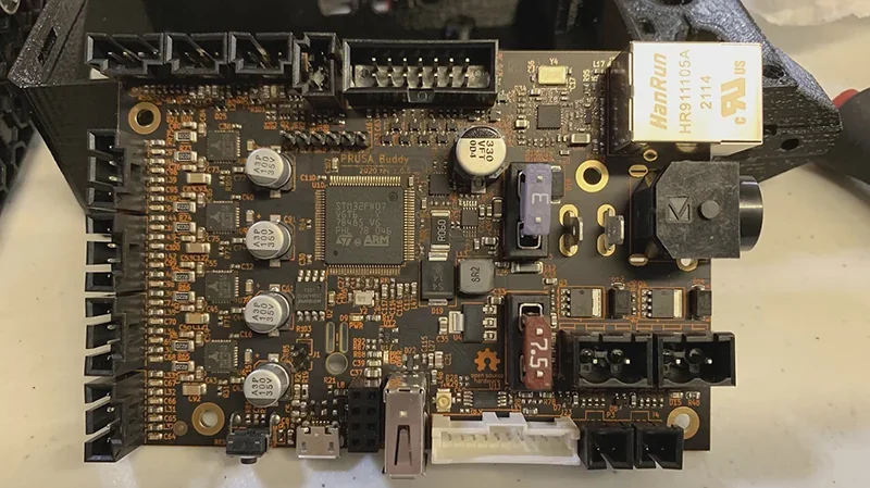 Prusa Mini 3D printer mainboard with capacitors, connectors, and circuits on a table. Branding visible, showing intricate electronic components layout.