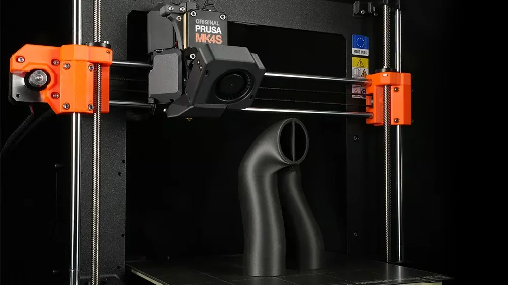 Prusa MK4 3D printer with black dual pipe print sample on build plate, featuring visible branding and orange components in a dark setting.