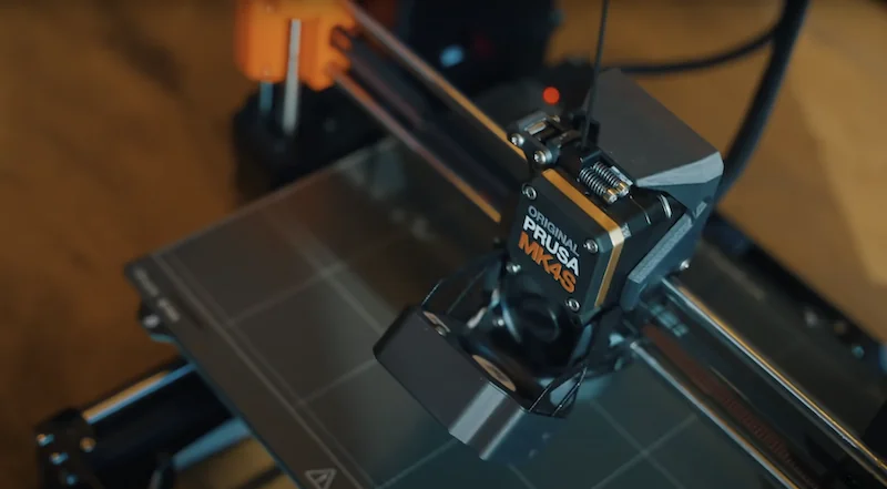 Prusa MK4 3D printer close-up showing the print head assembly and branding, positioned above the print bed in a workshop setting.