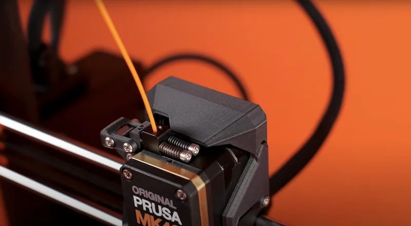 Prusa MK4 extruder with filament against an orange background, showcasing the 3D printer's sleek design and precise components.