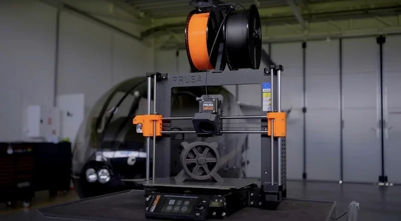 Prusa MK4 3D printer in a workshop setting, printing a gray wheel model with orange spool on top, featuring branding and touchscreen controls.