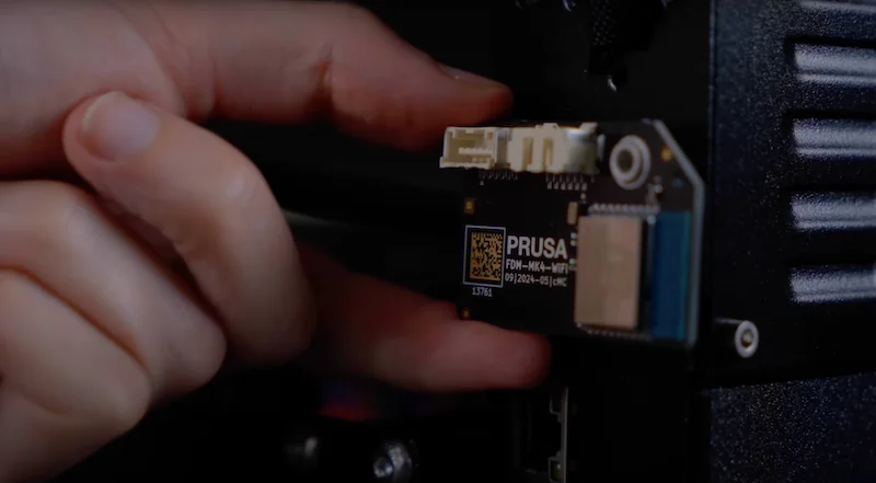 Prusa MK4 3D printer component installation close-up, focused on a hand connecting a Wi-Fi module, showcasing precise parts integration.