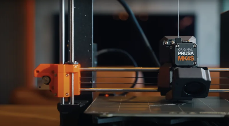 Prusa MK4 3D printer with orange and black components, showing detailed mechanics for precision printing. Branding visible.