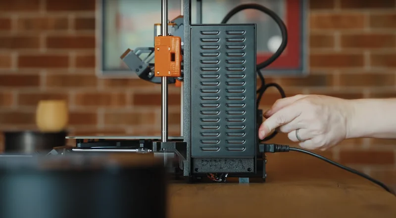 Prusa MK4 3D printer on a wooden table with a hand plugging it in, featuring visible components, against a brick wall backdrop.