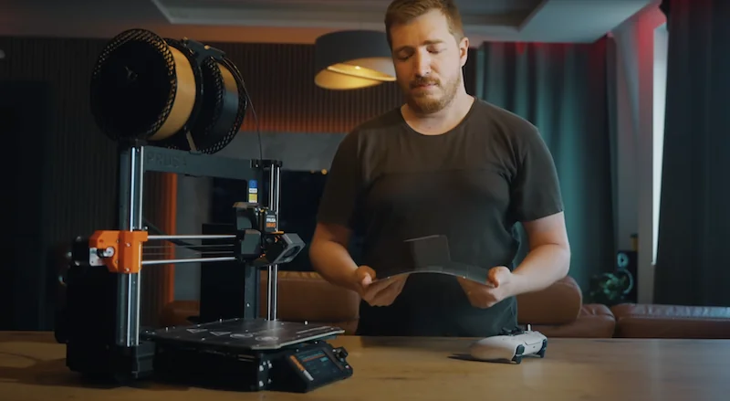 Prusa MK4 3D printer on a wooden table with a man holding a printed part, filament spool on top, in a modern room setting.