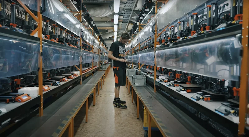 Prusa MK4 assembly line with worker inspecting 3D printers in a factory setting; rows of machines and parts produced by the Prusa MK4 visible.