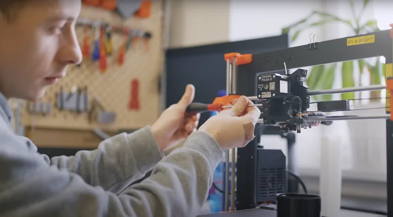 Person operating Prusa MK4 3D printer in workshop; tools and parts in background.