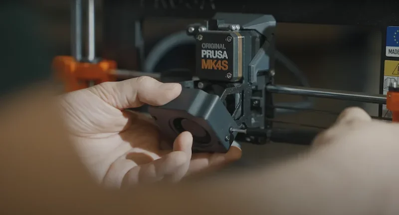 Hand adjusting Prusa MK4 3D printer component, showcasing brand logo and technical details.