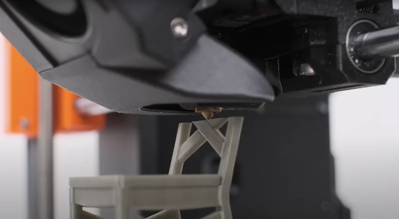 Prusa MK4 printing a small chair model with precision. Visible orange branding and nozzle close-up demonstrate 3D printing process.