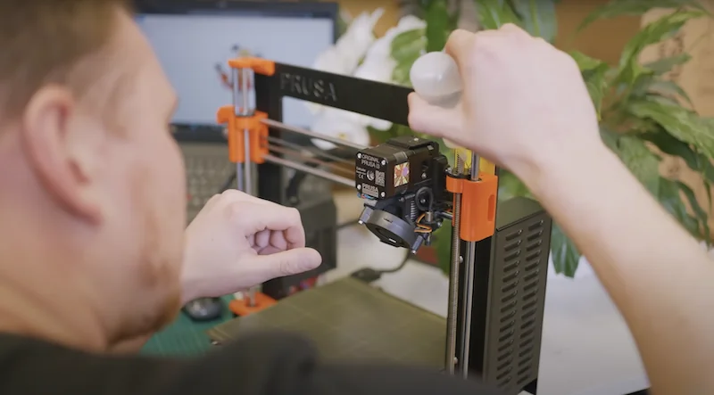 Prusa MK4 3D printer in use, with a person adjusting the extruder, showcasing the machine's design and features in a workspace with a laptop and plants.