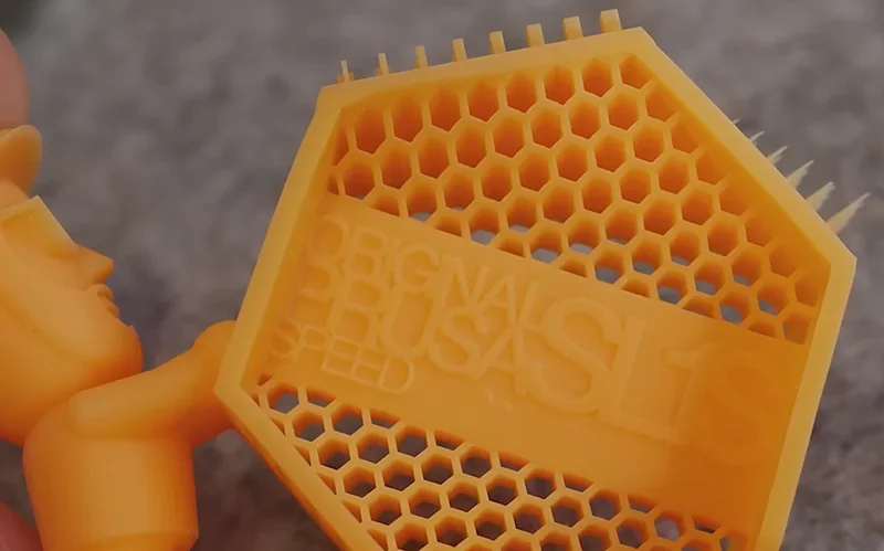 Prusa SL1S 3D printed hexagonal sample in yellow with honeycomb pattern, showcasing intricate detail and precision on a textured background.