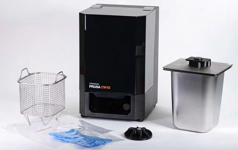 Prusa SL1S in black with a metal basket, stainless container, gloves, and parts on a white background.