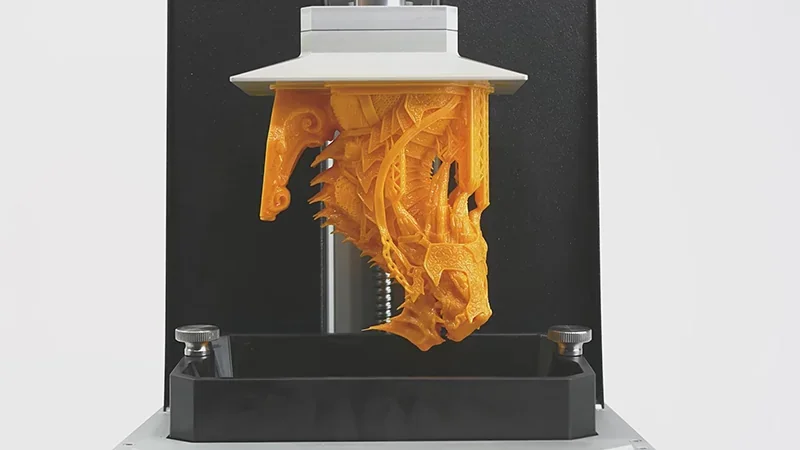 Prusa SL1S prints an intricate orange dragon model with precise detail, hanging from the 3D printer's build plate above a resin vat.