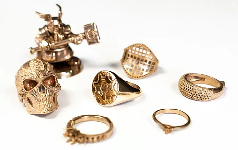 3D-printed gold rings and figurines crafted by Prusa SL1S, showcasing intricate designs, arranged on a white surface.
