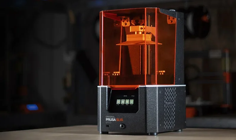 Prusa SL1S 3D printer displaying an orange-tinted cover and digital control panel, set in a workshop environment.