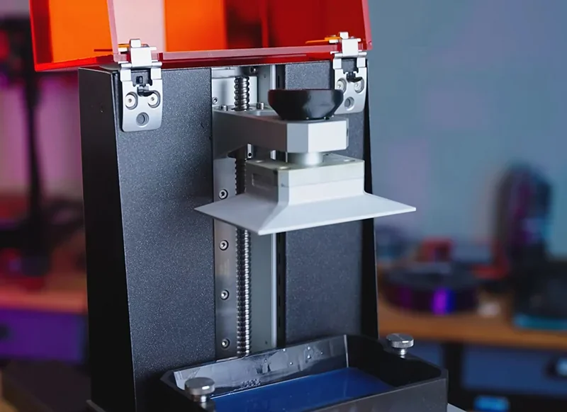 Prusa SL1S 3D printer with open orange cover, showcasing resin tank and build platform in a workshop setting.