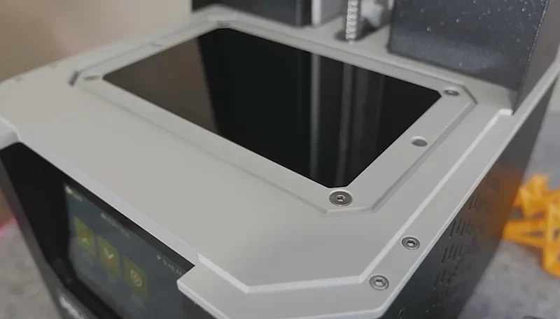 Prusa SL1S 3D printer top view showing sleek design with a touchscreen interface, featuring yellow printed samples on a gray surface.