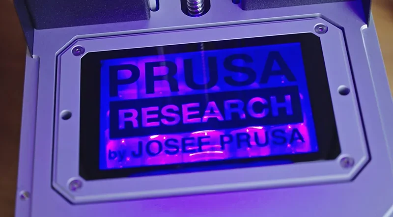 Prusa Research branding on Prusa SL1S 3D printer screen, illuminated in blue light, featuring robust industrial design and precise detailing.
