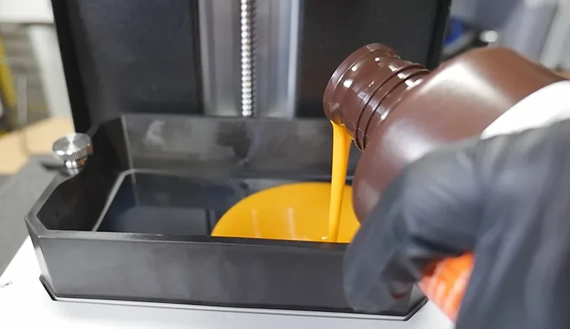 Pouring resin into a Prusa SL1S 3D printer's tray, gloved hand holding the bottle, ready for precision printing of detailed parts.