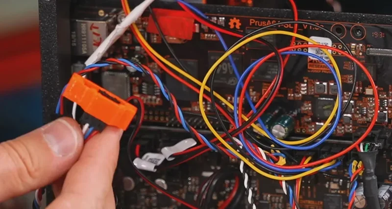 Close-up of Prusa SL1S 3D printer electronic components and wiring, with a hand holding orange-printed part likely made by Prusa SL1S.