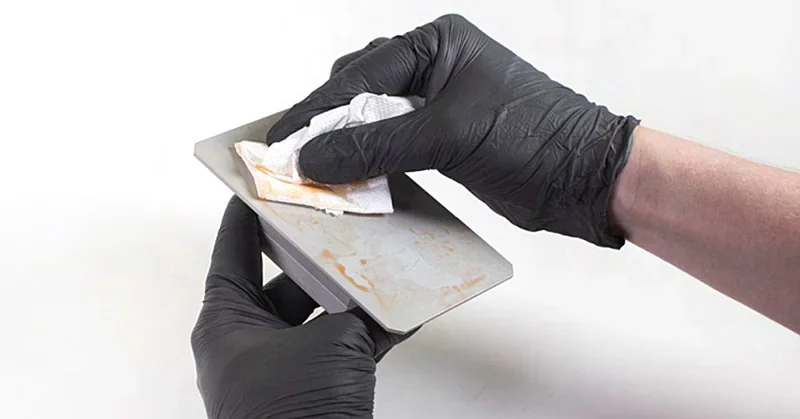 Cleaning 3D print plate with gloved hands, likely from Prusa SL1S printer. Wiping away residue on metal surface, ensuring smooth finish for accurate prints.
