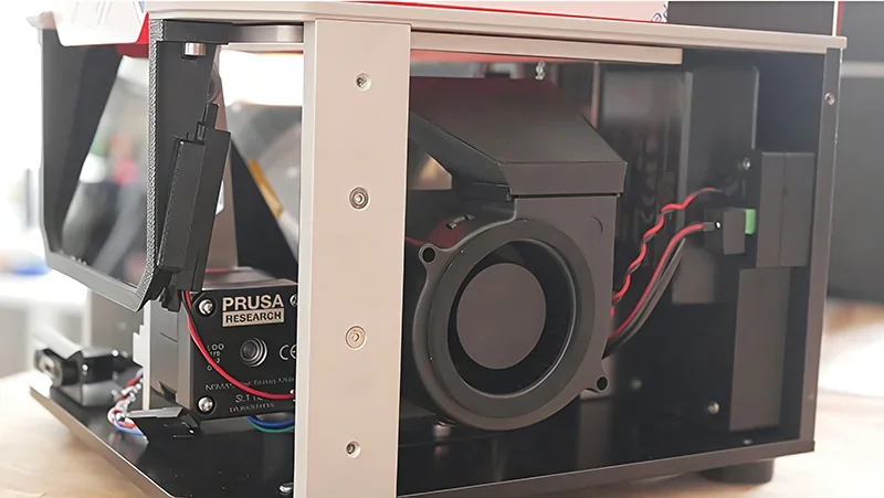 Prusa SL1S 3D printer interior with visible fan, wiring, and branding, on a tabletop setting, showcasing its technical components.